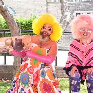 An Image Of Fay & Fluffy Outside, Sitting On Chairs. Fay Is Holding Up A Picture Book And Has A Yellow Wig, Pink Gloves, And A Floral Dress On. Fluffy Has A Pink Poncho, Leggings Lined With Cats, A Pink Wig And Pink Glasses. They Both Have A Large Smile On Their Face.