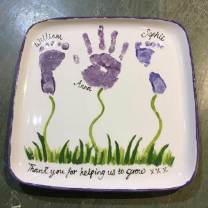 A White Square Plate With A Footprint, A Handprint, And Another Footprint Painted On It. The Prints Are In Purple, And Connect To Green, Painted Grass.