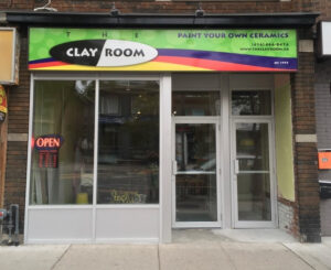  An image of the storefront of The Clay Room. The sign on top of the store reads “The Clay Room” in black and white, and “Paint your own Ceramics” in purple. The background of the sign is green, with strips of purple, orange and yellow.