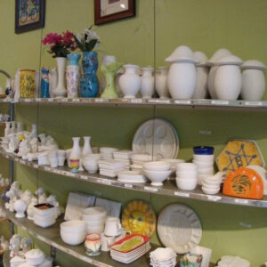 A Picture Of A Shelf In The Clay Room. The Shelves Display Many Kinds Of Ceramic Items To Be Painted. The Wall Colour Is Green-yellow.