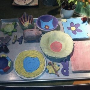 A Picture Of A Table Top With Different Ceramic Works In Progress. They Are Of Many Colours, And Include A Piggy Bank, A Turtle, Bowls And Plates.