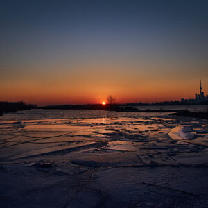 A Landscape Photo Of A Frozen Lake With The Sun Setting In The Horizon. The Sky Is A Gradient Of Blue To Orange. On The Right Side Of The Picture, We See The CN Tower And The Toronto Skyline.