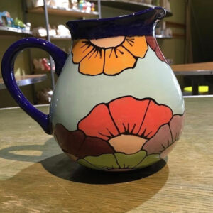 A Picture Of A Ceramic Jug, Painted In Light Blue, With Colourful Flowers. The Lip And Handle Of The Jug Is Deep Navy Blue.