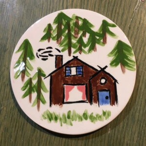 A Picture Of A Circular White Plate With A Brown House And Many Coniferous Trees Painted On It.