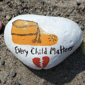 A Photo Of A Rock On A Sand Beach. The Rock Is Painted White, And Has An Illustration Of An Orange Moccasin Boot On It. It Reads The Words, 