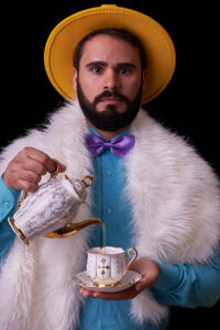 A portrait of Deivid. He has brown hair, with a beard and mustache. He is wearing a yellow brimmed hat, with a blue button up shirt, a purple bowtie, and a furry white scarf. He is holding an ornate teacup in his left hand, while pouring a liquid out of a teapot into it with his right hand. He is staring directly at the camera.