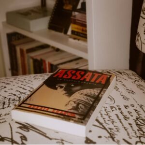 An Image Of A Book, “Assata: An Autobiography” By Assata Shakur, Placed On A Surface. In The Background Is A Blurred Bookshelf.