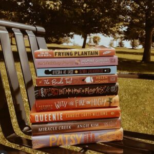 An Image Of A Stack Of 9 Books On A Park Bench. The Titles Include: Frying Plantain, The Wedding Party, Ayesha At Last, The Bride Test, Let Me Hear A Rhyme, With The Fire On High, Queenie, Miracle Creek, And Patsy.