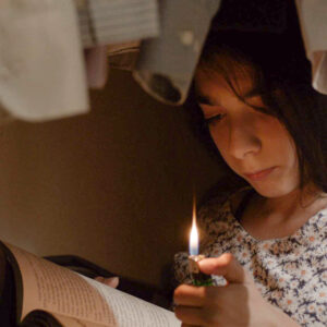 A Film Still Of A Young Child Holding A Lighter To Read A Book. She Appears To Be Under Hanging Clothes.