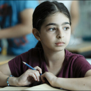 A Bright Film Still Of A Young Child In A Red T Shirt, Holding A Pencil And Sitting At A Desk. She Is Looking To The Side.