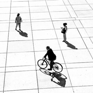 A Black And White Photograph Of A White Ground With Lines Running Through It To Make A Grid. The Shot Is Taken From A High Angle, Looking Down.There Are Three People On It: One Is On Bicycle, One Has Stopped And Is One Their One, And One Is Walking.