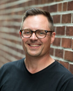 A headshot of Henry, with a black shirt and glasses on. He is smiling directly at the camera and standing in front of a brick wall.