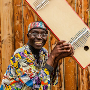 A headshot of Njacko Backo. He has dark skin and is smiling at the camera. He wears rectangular frame glasses, wears bright patterned clothing and is holding up a kalimba.