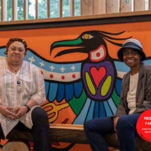 A photo of Carol and Bernnitta sitting in front of a bright mural by Indigenous artists Philip Cote. The mural is an Indigenous-style bird with wings spread wide. The person on the left has medium-dark skin and wears a striped top. The person on the right has dark skin, a blue bucket hat and a gray blazer. They are smiling.