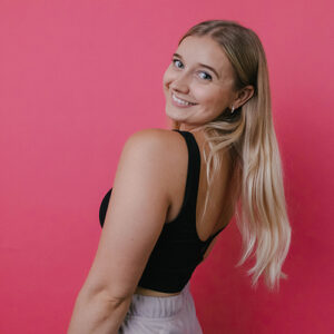 A headshot of millie bee. She has long blond hair, light skin, and is wearing a black sleeveless top. She’s smiling over her shoulder, standing in front of a bright pink backdrop.