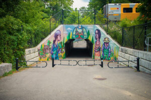 An exterior shot of a mural on the face of a tunnel. A walking path leads through the tunnel. The mural features four colourfully rendered people gardening together.