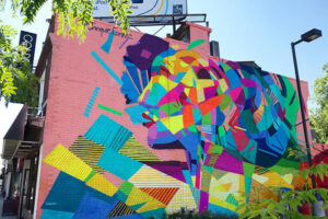 A picture of a mural that covers the entire side of a two-story building. Colourful abstract shapes form the impression of a woman's profile on a light pink background.