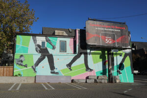 A picture of a mural. The mural takes up the full side of a two-story building. It features three figures in grayscale walking on a bright green and blue background.