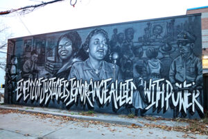 A picture of a grayscale mural, in bold letters it reads, "the enemy of justice is ignorance allied with power". At the centre of the mural is two women, one speaks into a microphone.