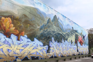 A picture of a massive mural on the side of a building. Across the bottom in blue and yellow are three graffiti style words. Behind them rises a realistically rendered Machu Pichu.