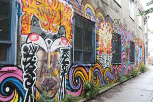 A picture of a mural along a long wall. The mural is chaotic and vibrant, featuring bright oranges, pinks and purples. There are several faces painted between windows, along the bottom are swirling waves with bold black outlines.