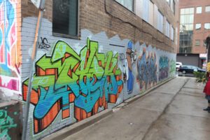 A photo of an alley way. Vibrant graffiti-style words cover the wall. Nearest to the camera is bright green and blue street art.