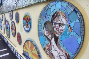 A picture of several round mosaics attached to a yellow painted wall. Feature a variety of scenes in blue and yellow tile, including a portrait and a sunrise.