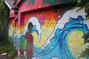 A picture of a mural painted on a garage. The mural is of a person with dark skin in a red bikini. Bright blue waves crash behind her on a vibrant yellow and orange background.