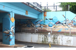 A picture of an underpass painted with a bright blue mural. Beige geometric shapes with bold black lines cut through the blue.