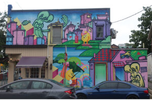 A picture of a pastel-coloured mural on the side of a building. At the front of the building is a storefront with a purple awning. The mural features stylized bunnies throughout a pink-tinted urban scene.