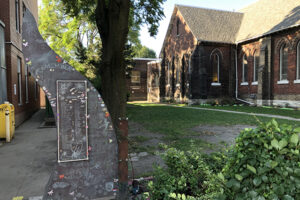 An exterior shot of a park with a brick church in the background. In the foreground is a metal sculpture with writing and drawing all over it in different handwriting.
