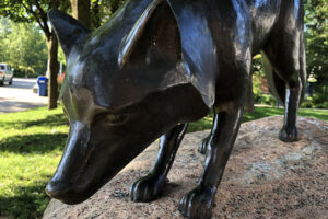 A picture of a bronze statue of a fox. The statue rests on a rock in a park.