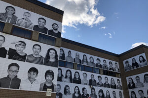 A picture of a side of a school. Across the walls are pasted 200 black and white portraits of students.