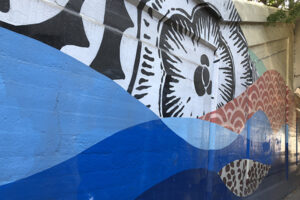 A picture of a mural on the side of an underpass. There are a blue waves and a red wave featuring a decorative pattern. Above the waves a black and white flower-type shape is painted in bold linework.