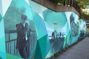 A picture of a mural on the side of an underpass. In prismatic blues there are several outdoor scenes painted. In one, a person leans against a metal railing drinking a coffee, in another there is a beach scene.