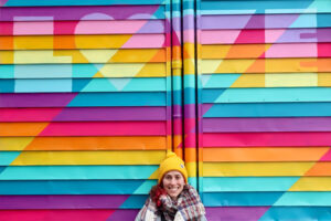 A picture of a woman in a yellow hat in front of a colourful mural. She is smiling. The mural is a bright rainbow and in lighter block leaders it reads, "Love".