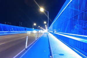 A picture of the Luminous Veil lit up a night. It is a bridge, bordering the edges is a wire wall lit up bright blue.