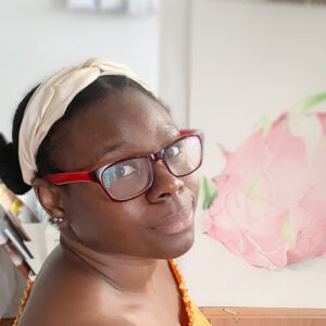 A headshot of Kendra. She has dark skin and wears red rectangle-framed glasses. She's sitting in front of one of her paintings and looking at the camera over her shoulder.