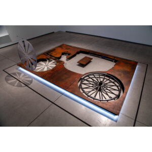A Photo Of An Art Installation. It Features A Sheet Of Rust-painted MDF With A Silhouette Of A Carriage Cut Out Of. One Of The Steel Spoked Wheels Stands Upright Perpendicular To The Rest Of The Installation.