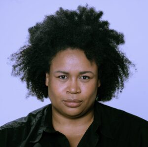 A headshot of Tracey Prehay. They have dark skin and poufy textured hair, they wear a black shirt and stare directly at the camera.