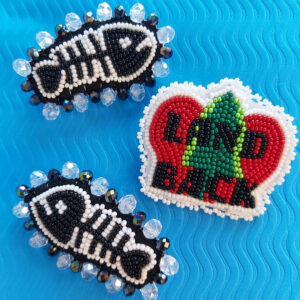 A Photo Of Hand-beaded Pins. Two Are Fish Skeletons, One Features A Three With 'LAND BACK' Beaded Across In Black.