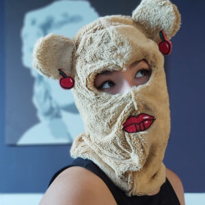 A Picture Of A Woman Wearing A Bear Balaclava With Red Lips And Cherry Earrings That Have Been Beaded Onto The Bear.