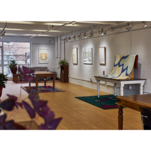 An Interior Shot Of The Riverdale Gallery. There Are Several Tables Displaying Textile Art. The White Walls Have Framed Art Hung Along Them.