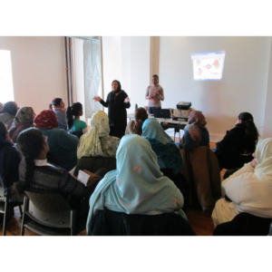 A Photo Of A Workshop Or Talk, A Woman In A Hijab Stands At The Head Of A Class Talking Animatedly.