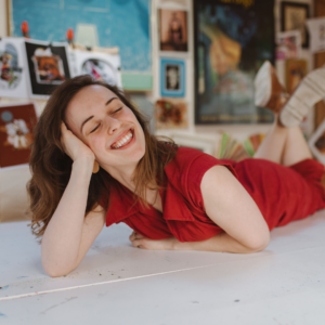 A photo of a woman laying on her stomach on a white table. She wears a red dress, has long brown hair and is smiling with her eyes closed.