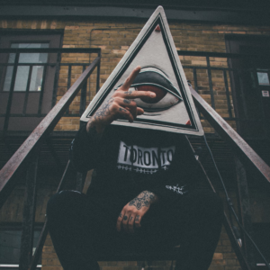 A photo of Flips. He's sitting on a fire escape and has a one-eyed pyramid for a head. His black hoodie reads: "Toronto" in block text.