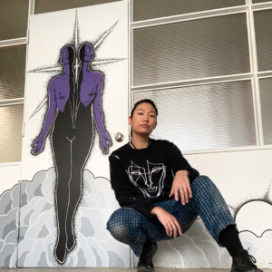 A Photo Of Victoria Day Crouched In Front Of A Mural. The Mural Is Of A Human Figure Whose Torso Is Split Down The Centre By A Star. Their Upper Half Is Opaque Purple And The Lower Half A Solid Black. The Figure's Hands Are At Their Sides, Palms Facing Outwards.