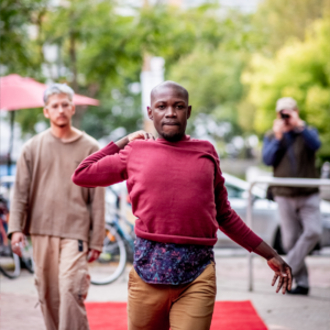 A Photo Of Pulga Moving Towards The Camera. They Are Wearing A Red Long Sleeve Shirt And Tan Pants. Behind Them Is Roberto, Out Of Focus And Also Facing The Camera.