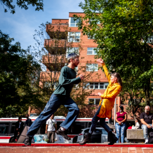 An Outdoor Shot Of Roberto Striding Towards Heidi, Who Steps Backward Across A Red Carpet. They Are Outside, Behind Them A TTC Bus Passes By.