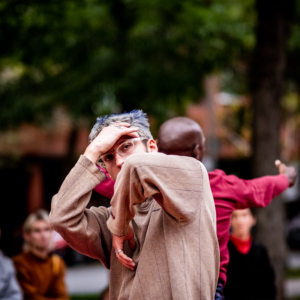 A Photo Of Roberto, He Is Dancing, Arms Crossed In Front Of Him. He Peers At The Camera Over One Arm, The Other Hand To His Forehead. Behind Him, Out Of Focus, Is Pulga In A Red Shirt.